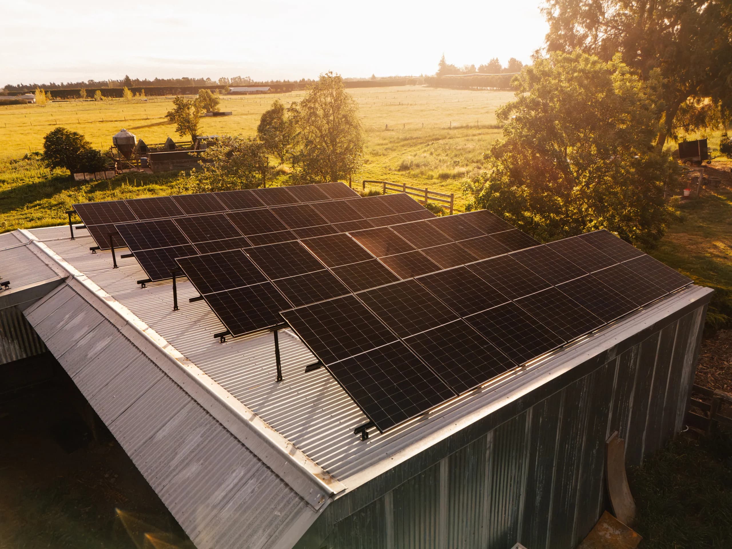 Close aerial view of a solar panel array on a corrugated shed roof lit by warm low sun with farmland beyond.