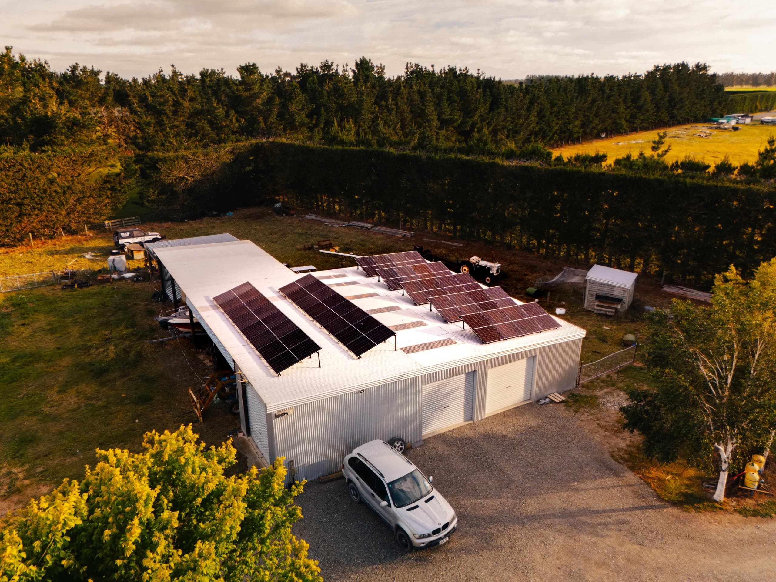 Drone view of a white farm shed with rooftop solar panels and a silver BMW SUV parked out front, surrounded by paddocks and pine shelter belts.