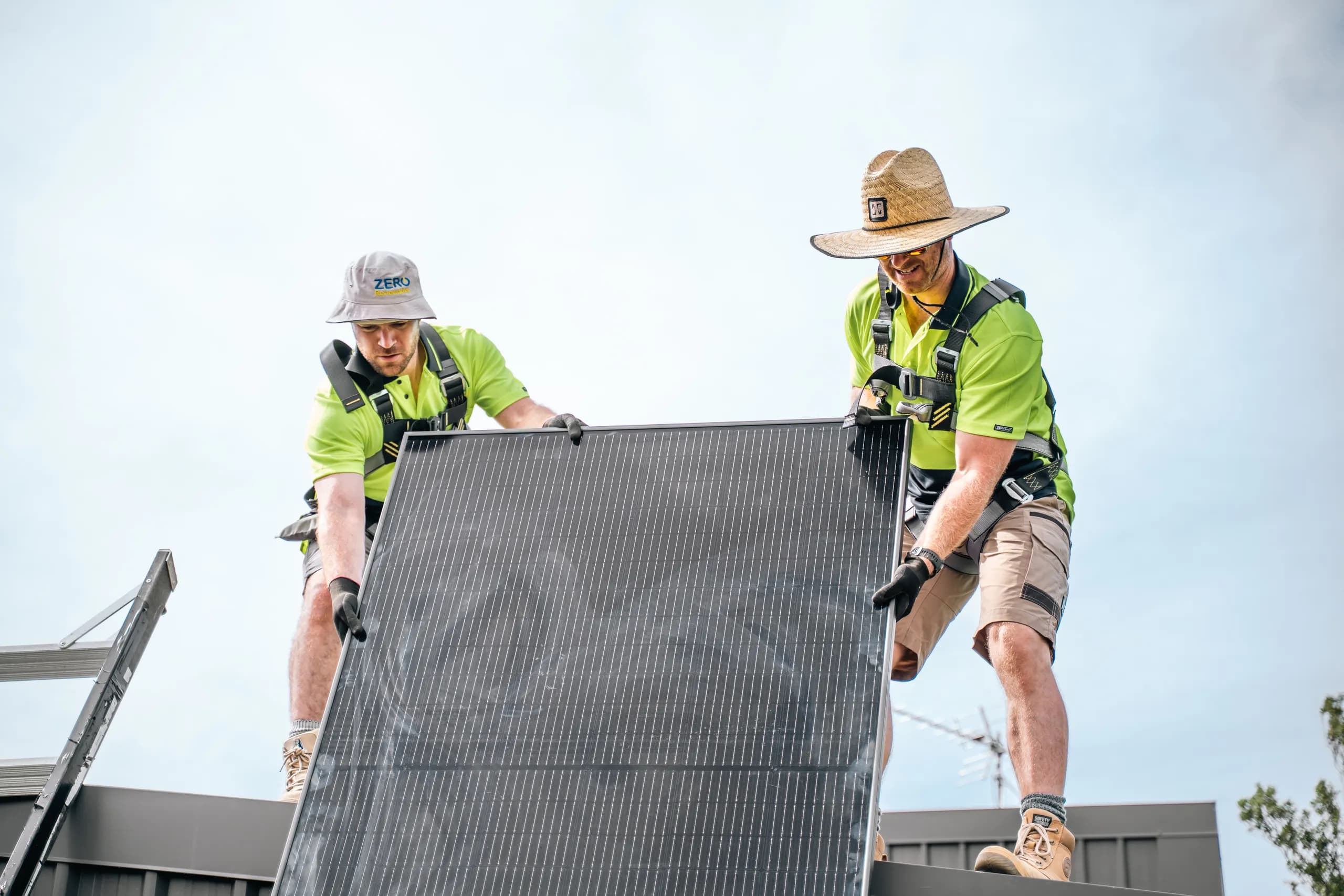 Two ZERO electricians in hi-vis and safety harnesses lift a solar panel onto a roof against a bright sky, one wearing a ZERO branded bucket hat.