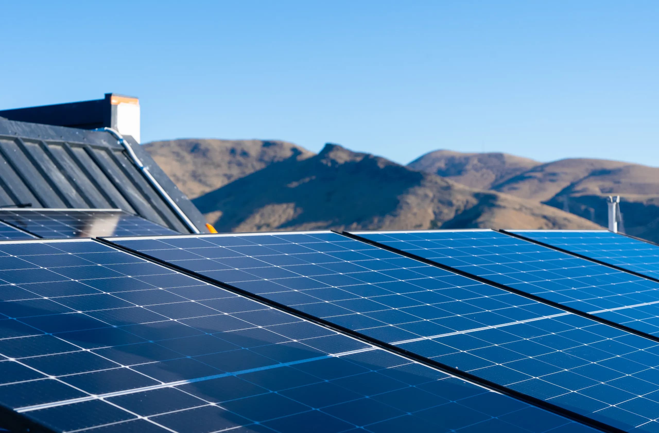 Close-up of blue monocrystalline solar panels on a metal roof with the Port Hills as a backdrop under a clear sky.