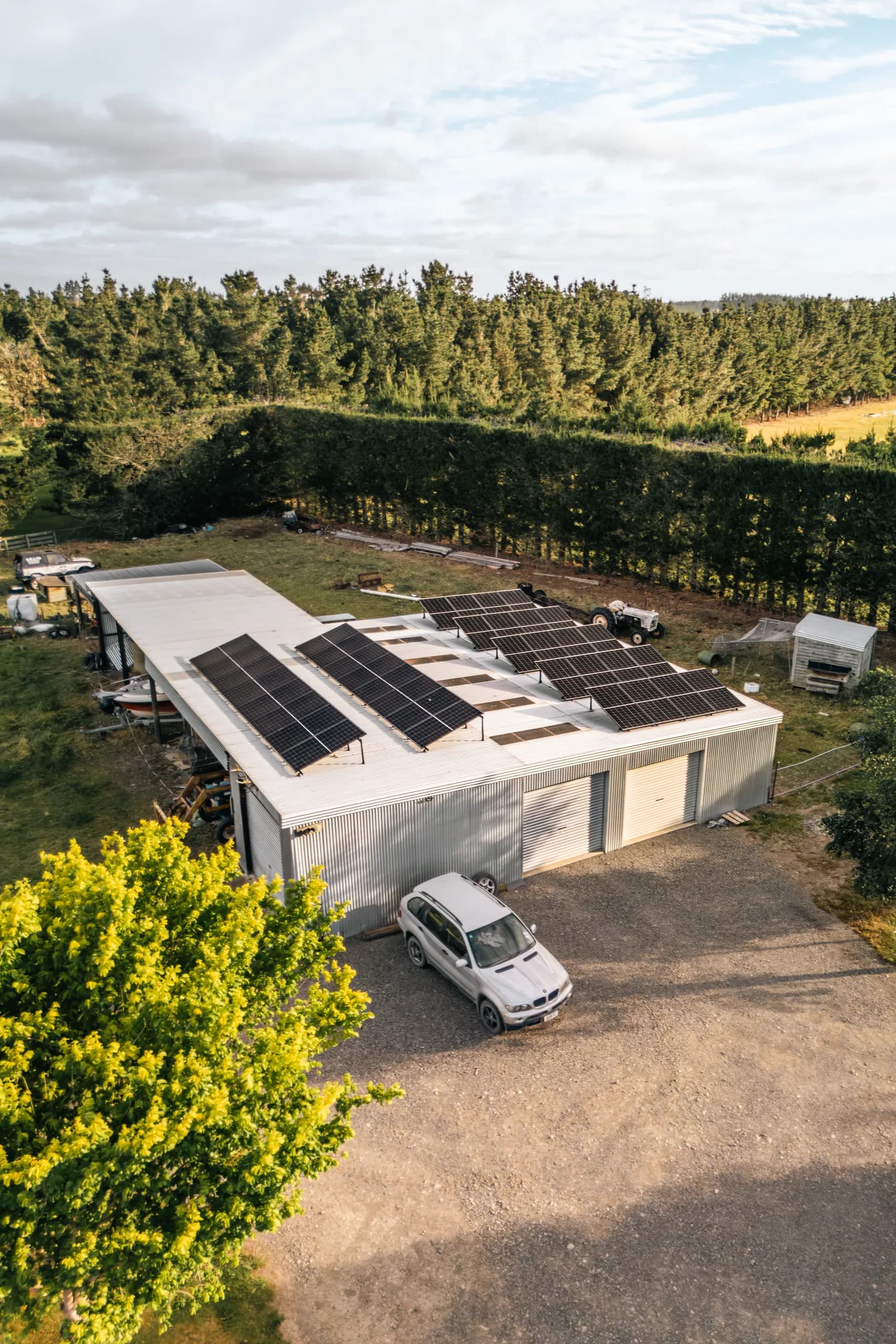 Close aerial view of a black solar panel array angled on a white shed roof with a driveway, parked SUV and green paddock and shelterbelt behind, shot in warm evening light.