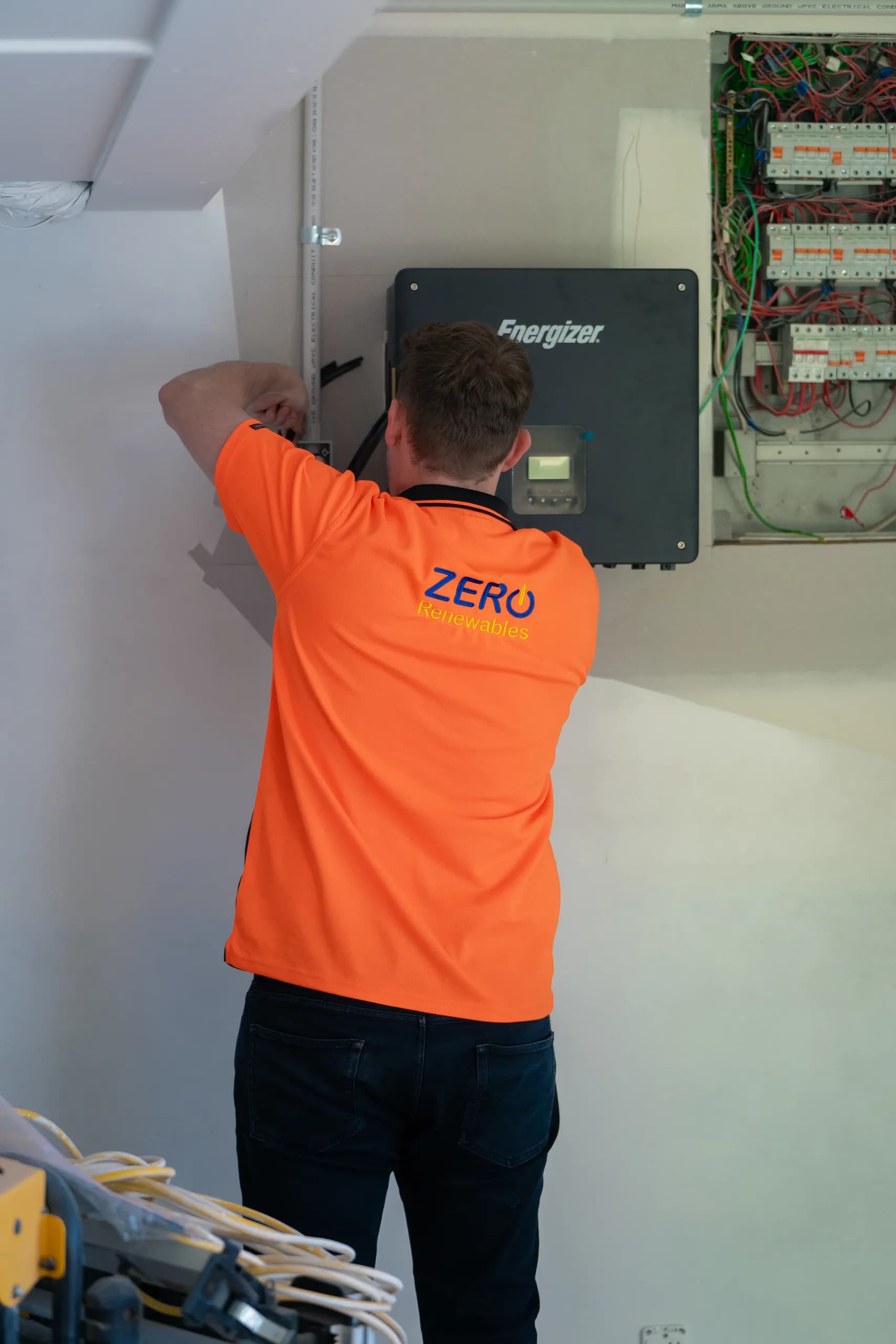 Technician in a Zero Renewables hi-vis shirt working on an Energizer hybrid inverter beside an open switchboard with exposed wiring.