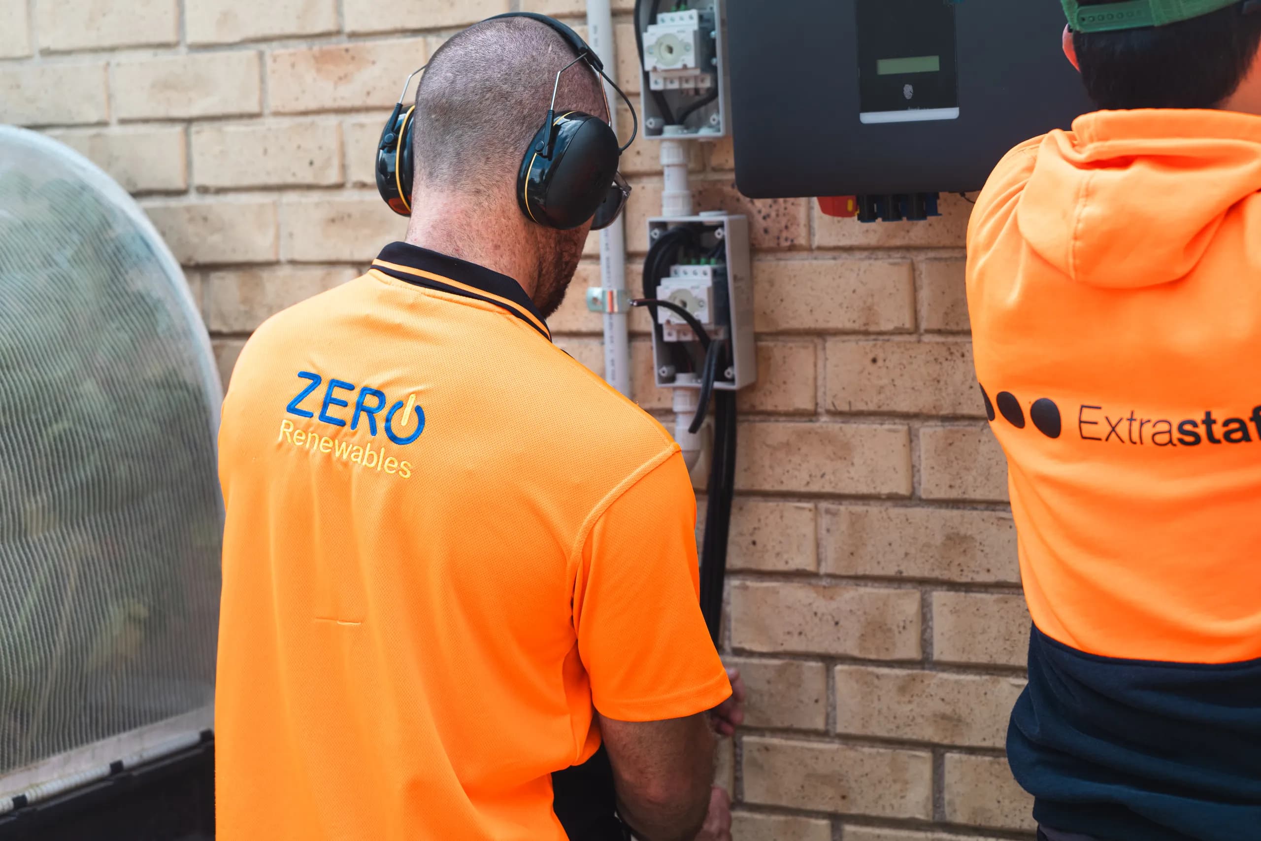 Zero Renewables technician in hi-vis and ear muffs working on wall-mounted isolators and an inverter, with a second worker in Extrastaff hi-vis beside him.