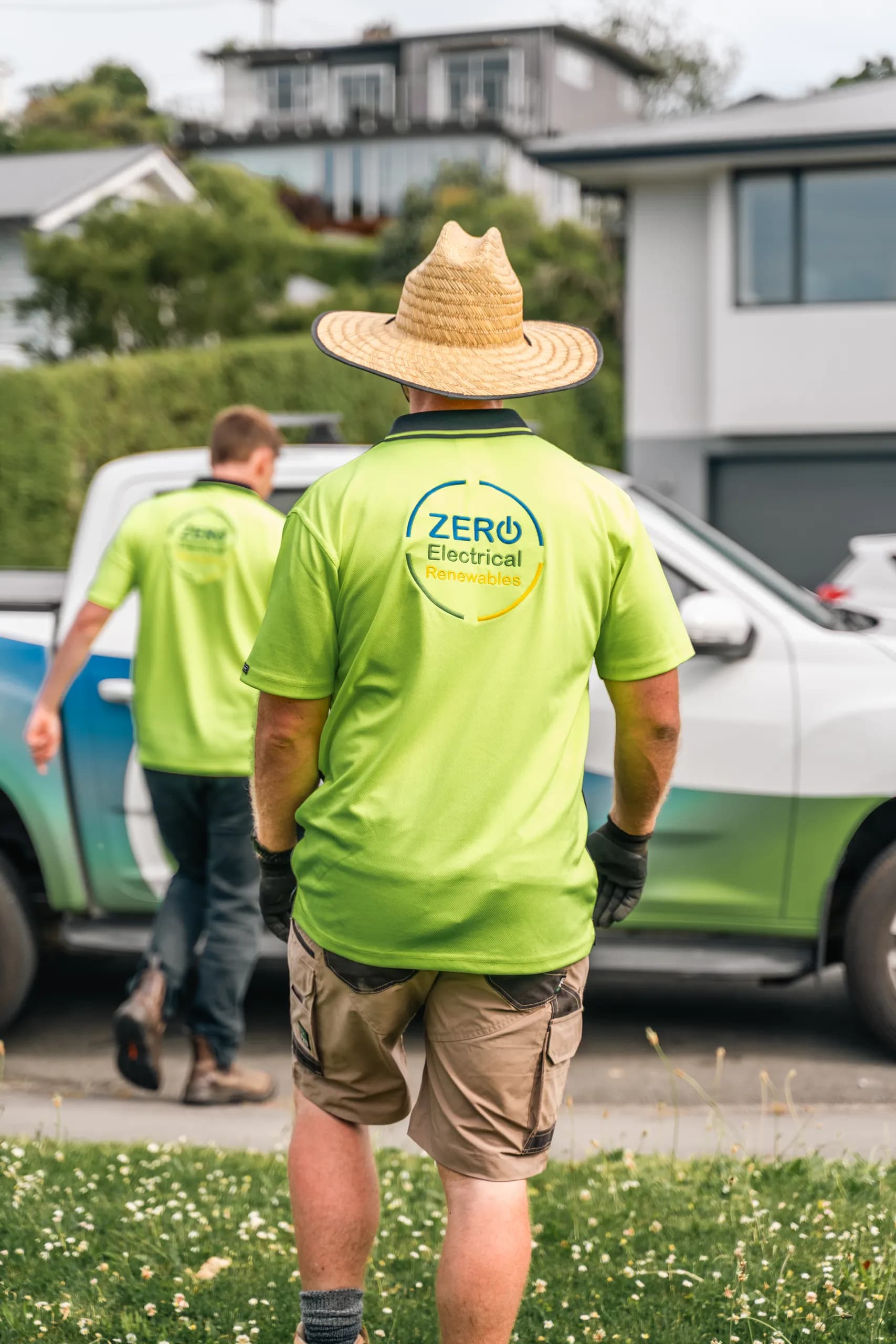 Rear view of two ZERO team members in branded hi-vis polos walking toward the company ute, with the ZERO Electrical Renewables logo on the shirt back.