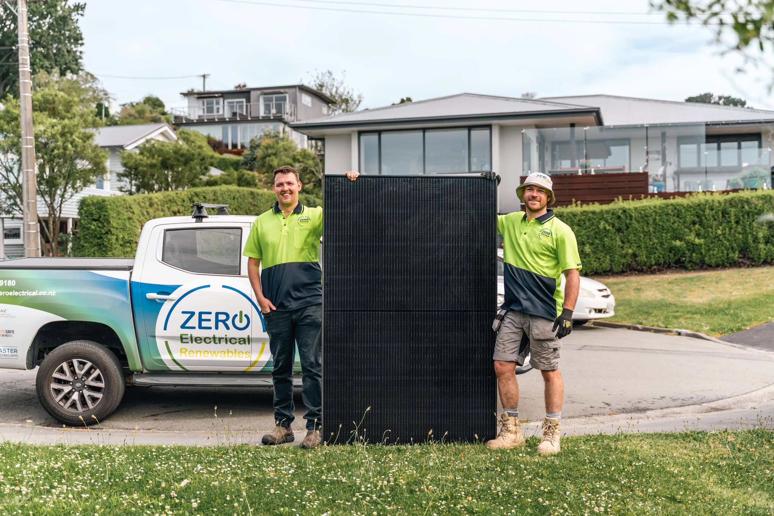 Two smiling ZERO electricians stand either side of a solar panel in front of the branded ZERO ute on a suburban grass verge.