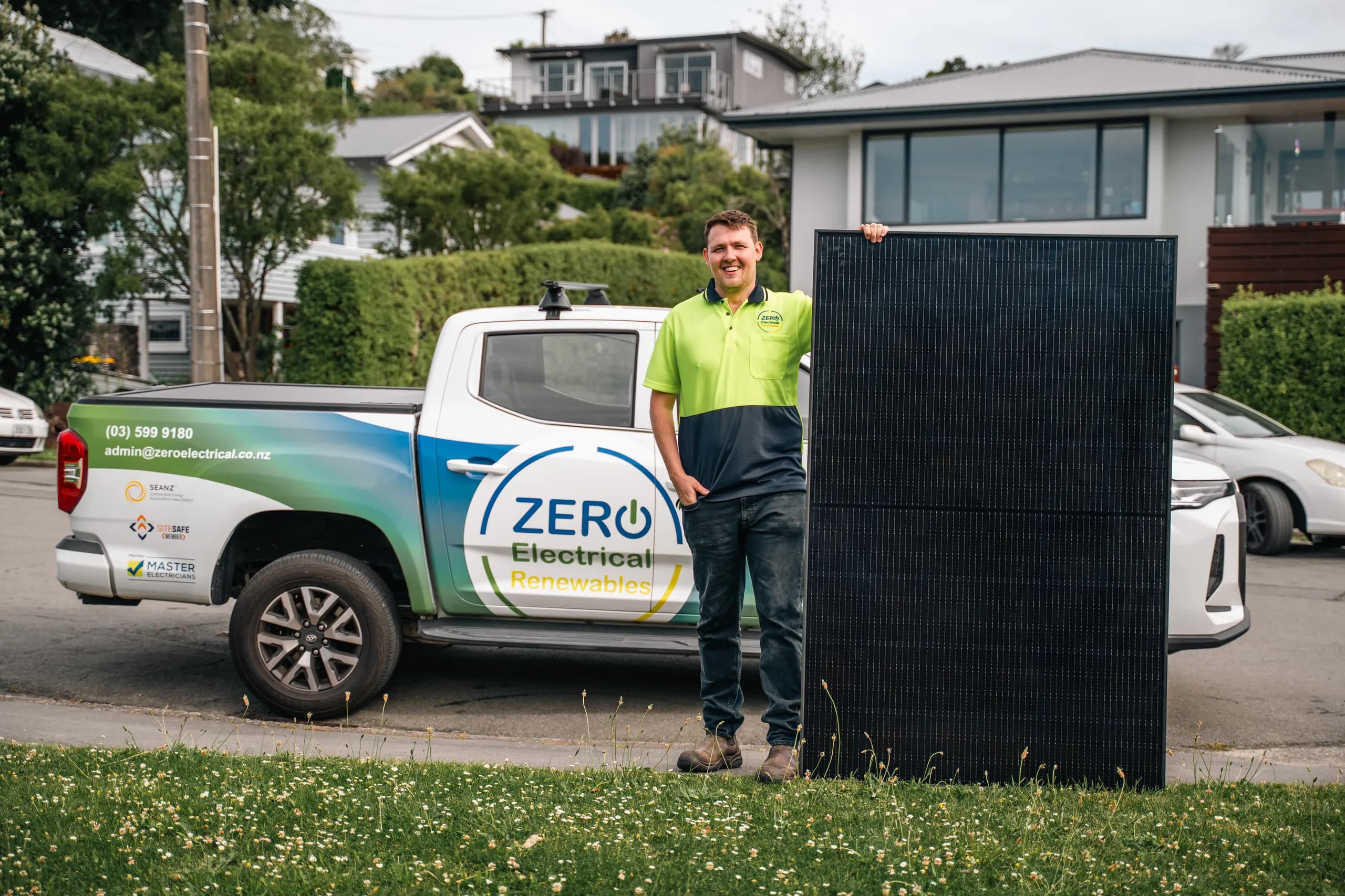 A smiling ZERO team member in hi-vis stands with a solar panel beside the fully branded ZERO Electrical Renewables ute on a suburban street.